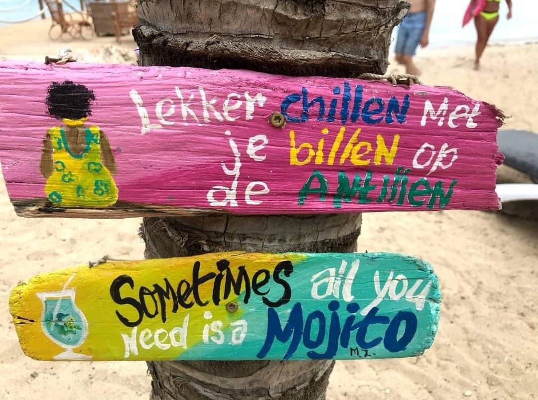 Kleurrijke houten bordjes op een strand op Curaçao met de teksten Lekker chillen met je billen op de Antillen en Sometimes all you need is a Mojito